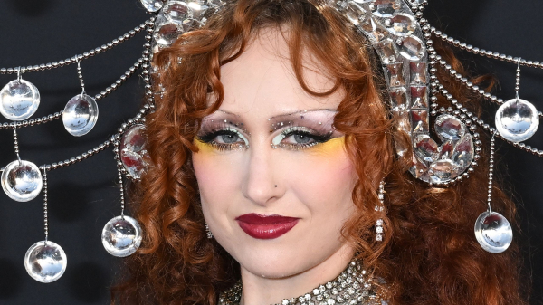 Chappell Roan poses at an event. She wears a beaded headdress and silver sparkling top. Her lipstick is red and she has...